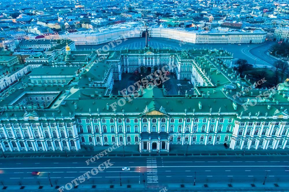На переднем плане изображён Зимний дворец — главное здание Государственного Эрмитажа, выполненное в стиле елизаветинского барокко. За ним раскинулась Дворцовая площадь с Александровской колонной и полукруглым фасадом здания Главного штаба. Композиция подчёркивает масштаб и симметрию исторического центра Петербурга. Вид с воздуха позволяет оценить планировку и архитектуру ансамбля. Снимок сделан ранним утром, когда улицы ещё пусты.